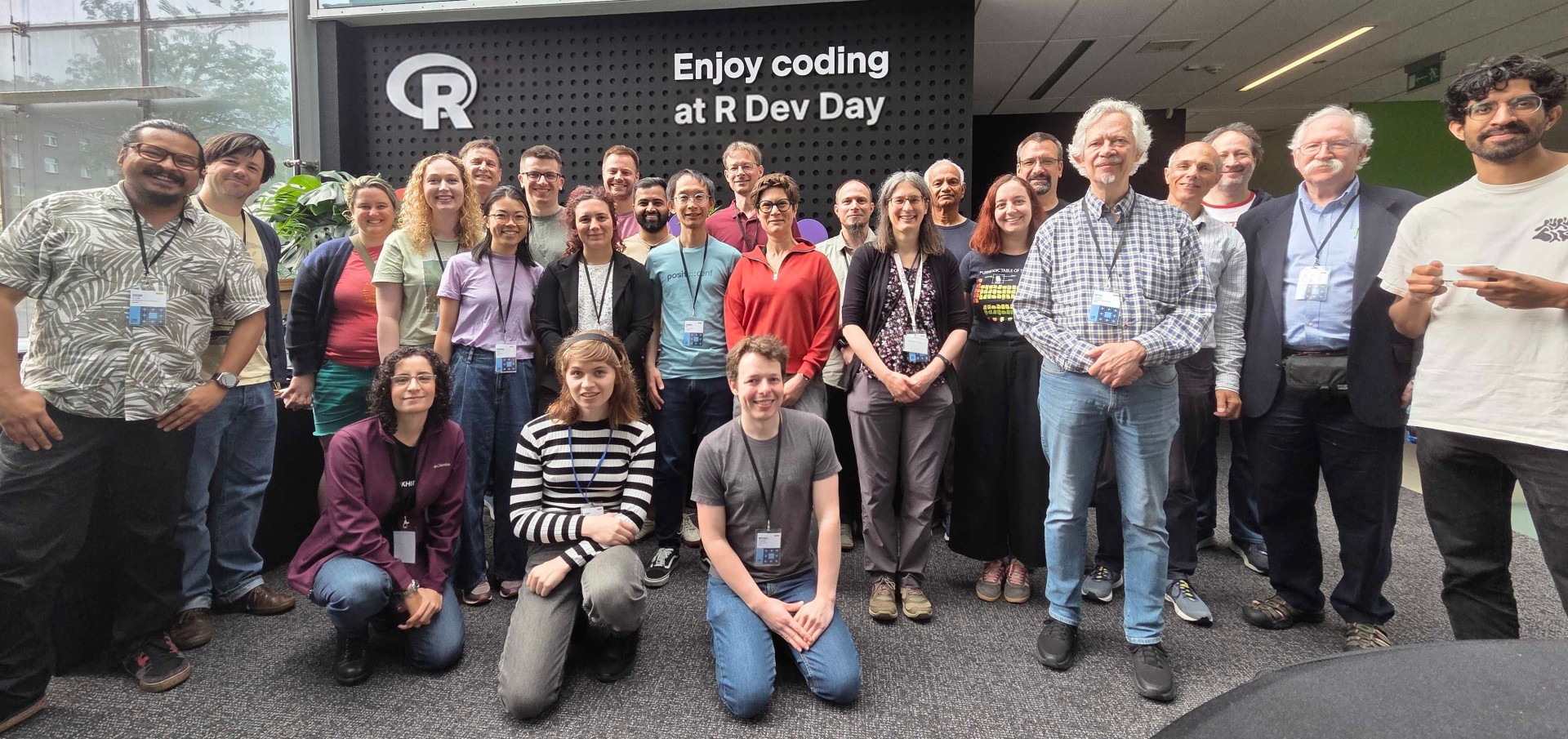 Group photo of R Dev Day 2026 participants in front of peg board wall with R logo and text 'Enjoy coding at R Dev Day'. There are 26 participants in the photo, with the author Mitchell O'Hara-Wild kneeling in the center of the front row.